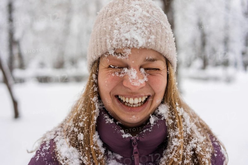 A young woman joyfully laughing, covered in snowflakes. She wears a purple jacket and a knitted hat, embodying the cheerful spirit of a snowy winter day