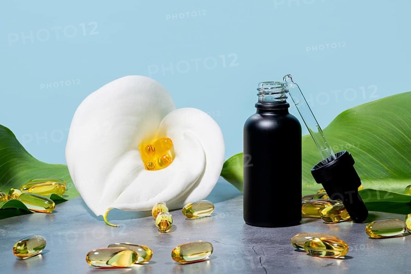 Mat black bottle of cosmetic serum next to a white white calla lily, featuring a pipette and scattered golden supplements omega capsules on a soft blue background