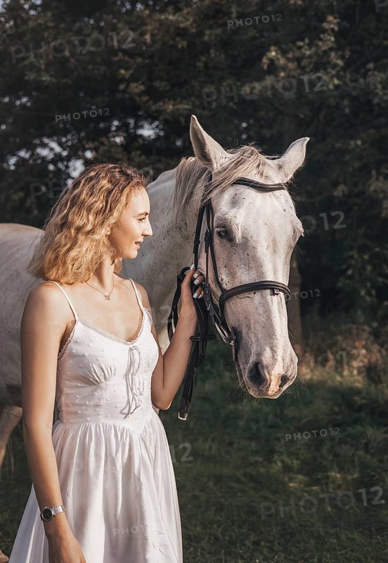 A young woman in a white dress stands beside a white horse, holding its bridle in a sunny meadow. The scene captures serenity and a connection with nature