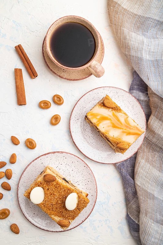 Honey cake with milk cream, caramel, almonds and a cup of coffee on a white concrete background and linen textile. Top view, flat lay, close up