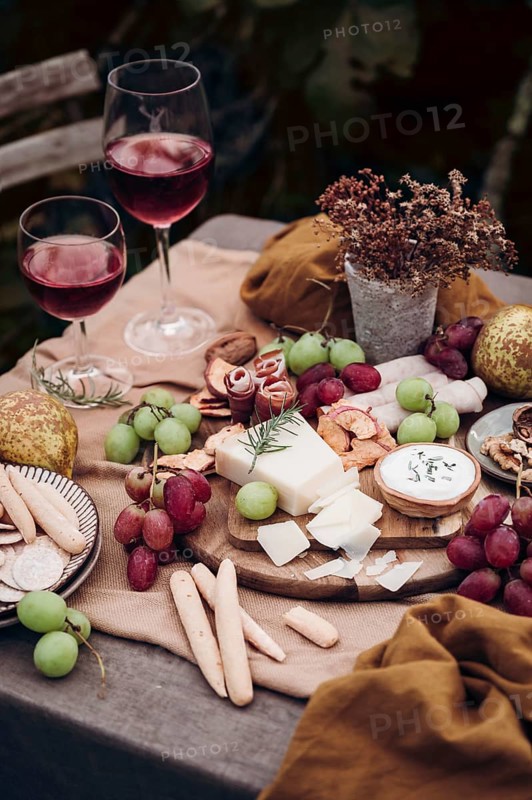 An elegant setting with a cheese board, assorted grapes, breadsticks, and cold cuts. Two glasses of red wine complement the gourmet snack arrangement, creating a perfect ambiance