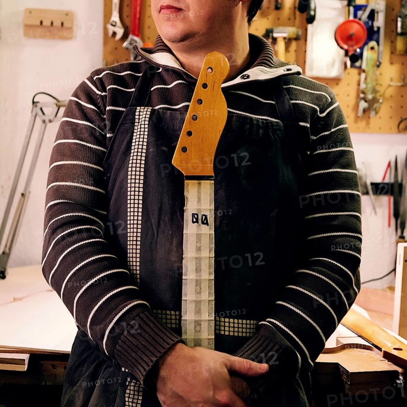 A luthier in a striped sweater and apron holds a bass guitar neck at their workshop. Tools are visible on the wall, showcasing a creative musical workspace