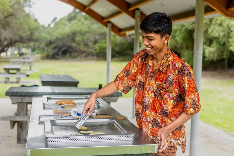 An Indonesian man wearing vibrant batik clothing enjoys cooking at an outdoor barbecue in Australia. Surrounded by green nature, he smiles while grilling food