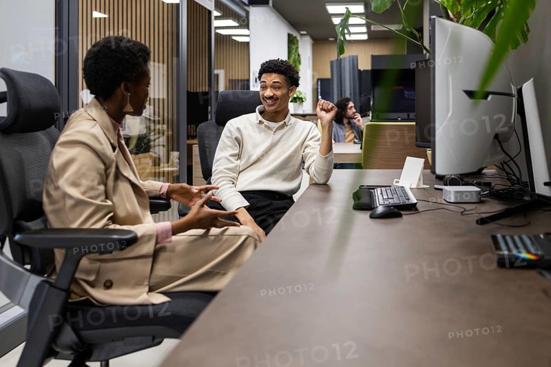 Two colleagues converse in a modern office setting, sitting across from each other. The environment is professional yet relaxed, with plants adding a touch of nature