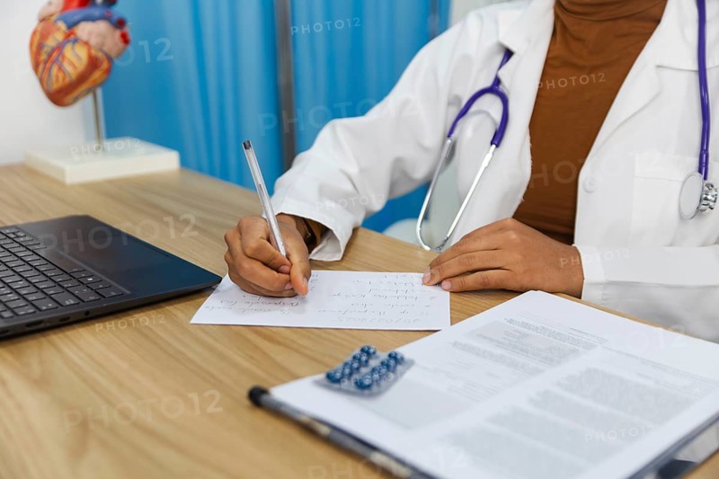 A doctor is seen writing notes on paper in a medical office. The scene includes a laptop, clipboard, and a stethoscope, representing a professional healthcare environment