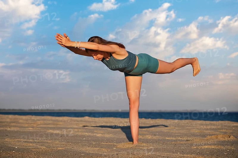 A woman gracefully performs the Virabhadrasana Warrior 3 pose on a sandy beach as the sun sets in the background. The calm waters and colorful sky create a peaceful atmosphere for yoga practice