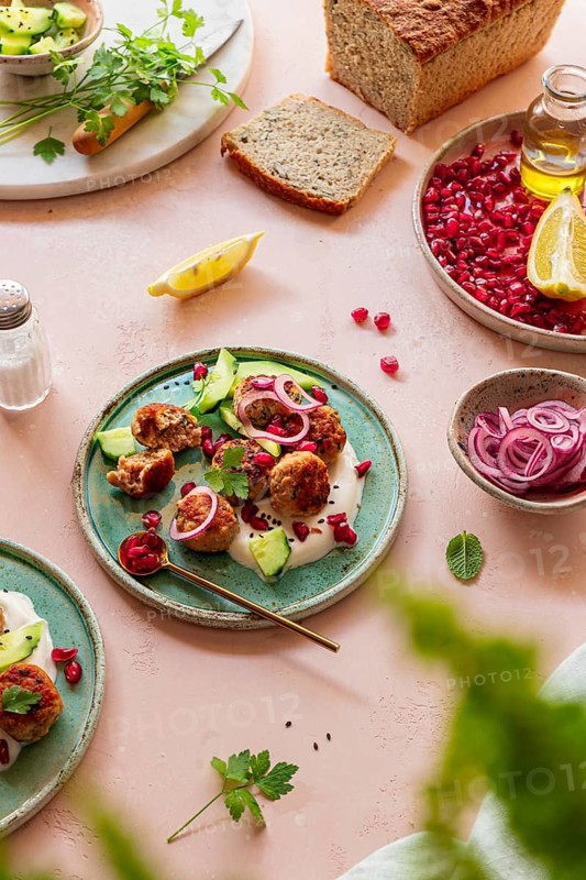 A vibrant Mediterranean meal featuring meatballs, pomegranate seeds, yogurt, sliced cucumbers, and pickled onions on a turquoise plate, accompanied by fresh bread and lemon wedges