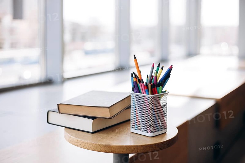 Colorful stationery items such as pencils and pens in a mesh holder alongside two hardcover books on a round wooden table, bathed in natural sunlight from large windows