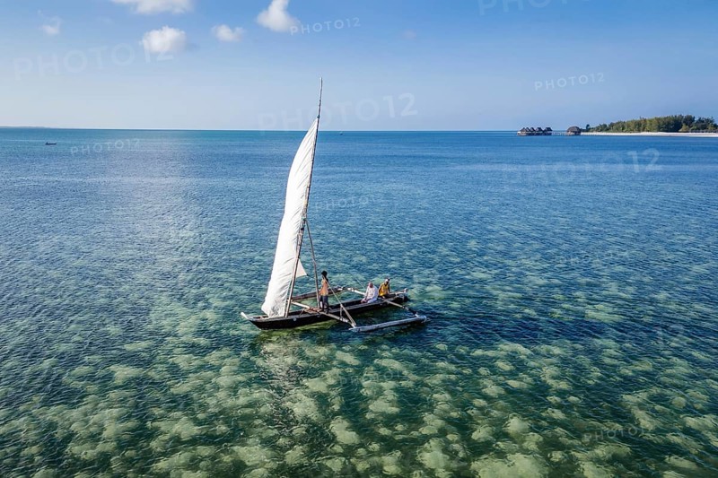 MICHAMVI KAE, ZANZIBAR - OCTOBER 14, 2025: High angle view of people sailing a traditional white-sailed ngalawa outrigger canoe over a clear coral reef in turquoise ocean water