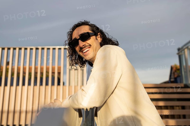 A fashionable man with sunglasses and a mustache relaxes on a sunny outdoor terrace His casual style and confident smile reflect a modern lifestyle vibe