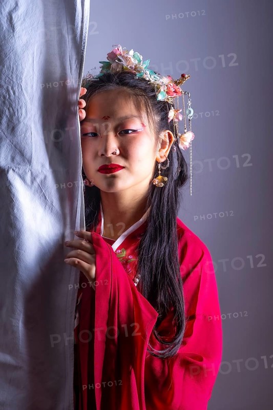 A young girl in a red dress with a flower headband stands behind a curtain. She has red lipstick on and her eyes are closed. Concept of mystery and intrigue