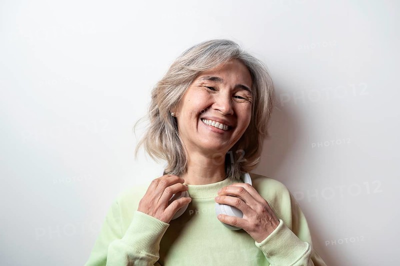 Asian senior woman with grey hair smiling joyfully, wearing green sweater and headphones, symbolizing music enjoyment and positive aging against a plain background