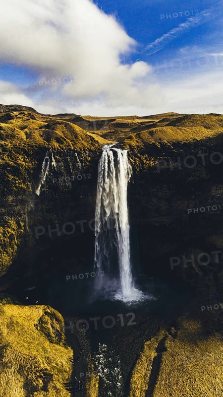 Striking aerial view of Seljalandsfoss waterfall in Iceland, cascading from a tall cliff surrounded by golden hills, under a vivid blue sky with soft clouds drifting above