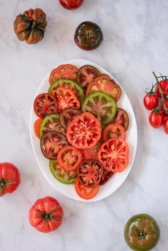 A variety of colorful heirloom tomatoes, sliced and arranged on a white plate, showcasing their vibrant red, green, and brown hues. Whole tomatoes surround the dish on a marble surface