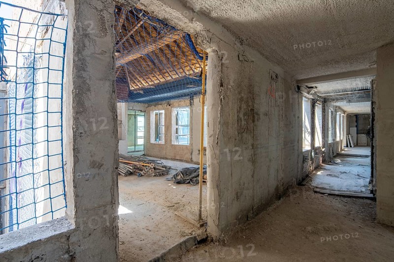 Interior view of a construction site showing exposed concrete walls and building materials. Sunlight filters through windows, illuminating the work in progress