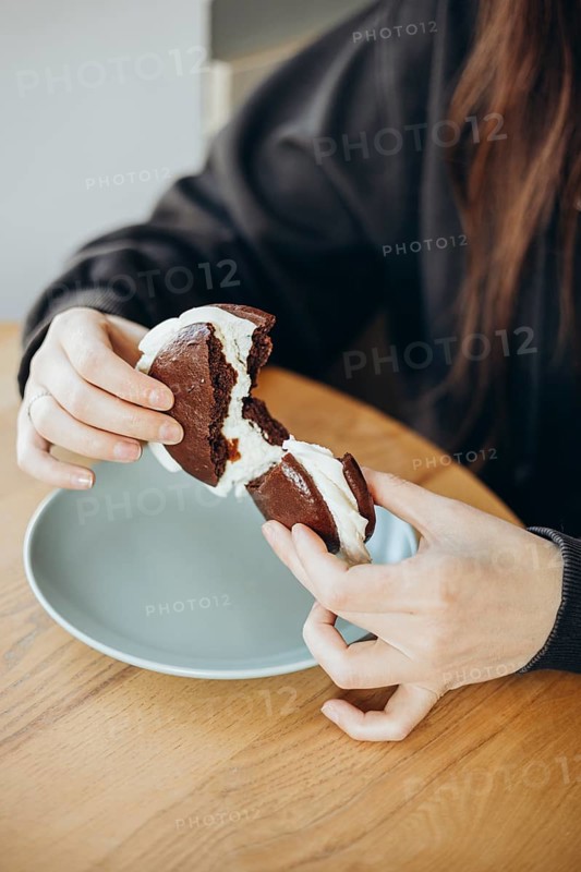 Close-up of a pair of hands pulling apart a whoopie pie, showcasing its creamy filling. The dessert sits on a light blue plate, offering a tempting look at the sweet treat