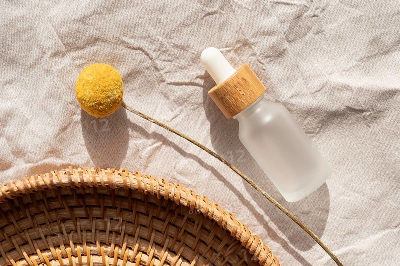 A minimalist scene featuring a cosmetic with a wooden cap on a textured fabric, accompanied by a yellow billy button flower and a woven basket, highlighting natural elegance