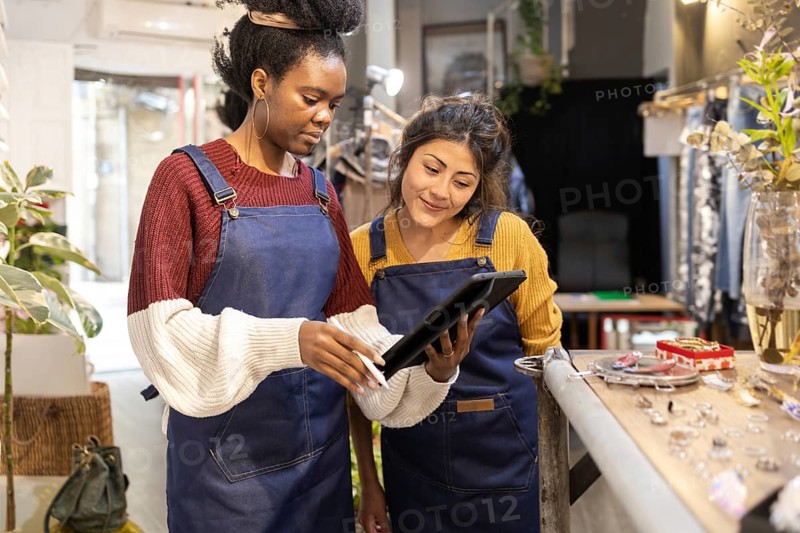 Two women in a fashion retail store, wearing blue aprons, collaborate using a digital tablet. They are surrounded by accessories and plants, showcasing teamwork and technology