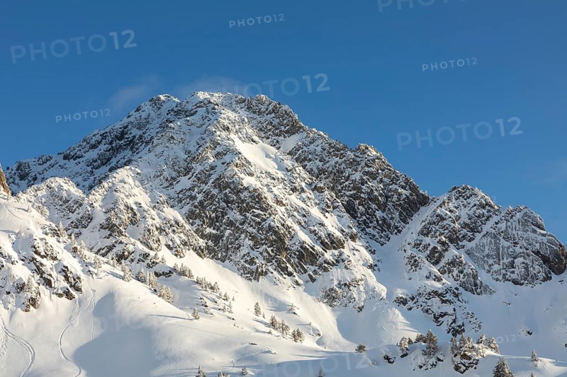 Stunning view of rugged, snow-covered mountain peaks illuminated by bright sunlight against a clear blue sky, highlighting the natural beauty and serenity of a winter landscape