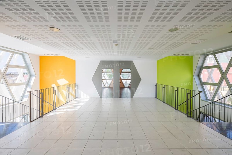 A modern auditorium hallway features hexagonal windows and vibrant yellow and green walls. The geometric ceiling design and sleek railing contribute to a fresh, contemporary ambiance
