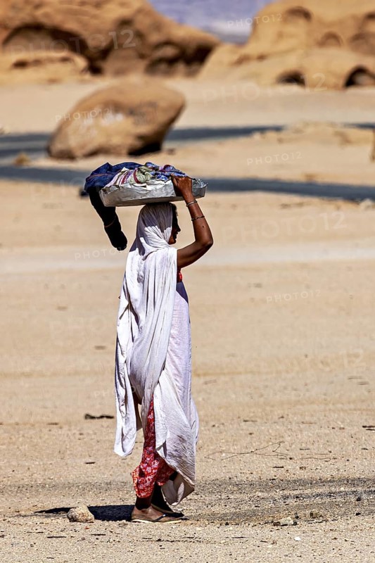 Woman in traditional dress carrying objects on her head, surrounded by a desert-like landscape, woman washing clothes in the Sahara in Algeria