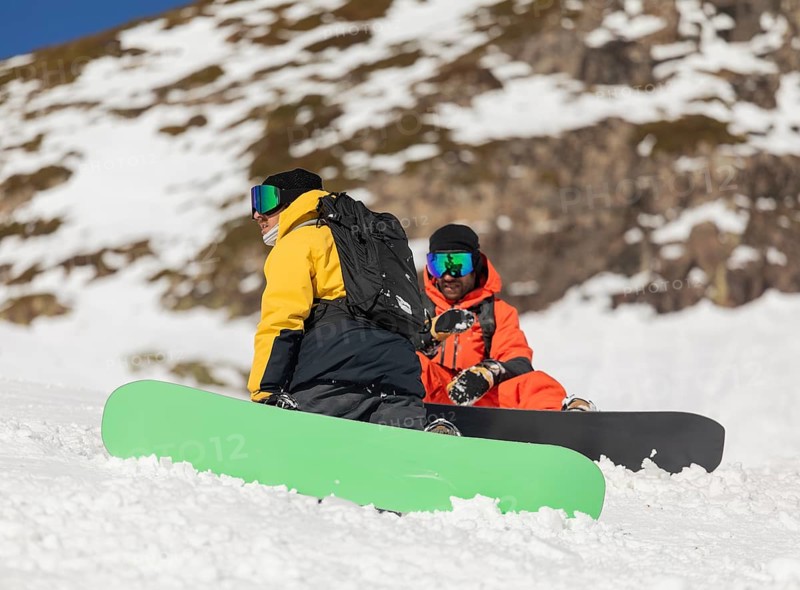 Two snowboarder friends, dressed in colorful winter gear, sit on a snowy mountain slope, surrounded by a scenic winter landscape, enjoying a break from snowboarding