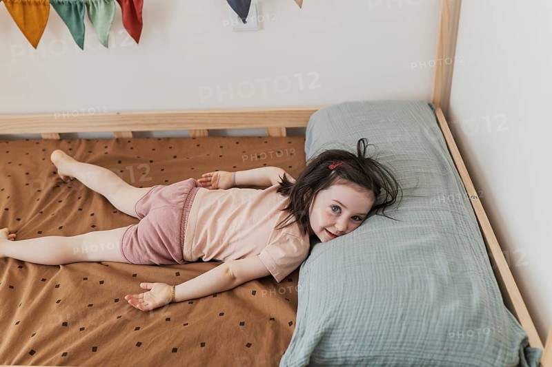 A cheerful young girl with dark hair and a playful expression lies on a cozy bed with geometric-patterned sheets and a large pillow, creating a warm and joyful scene