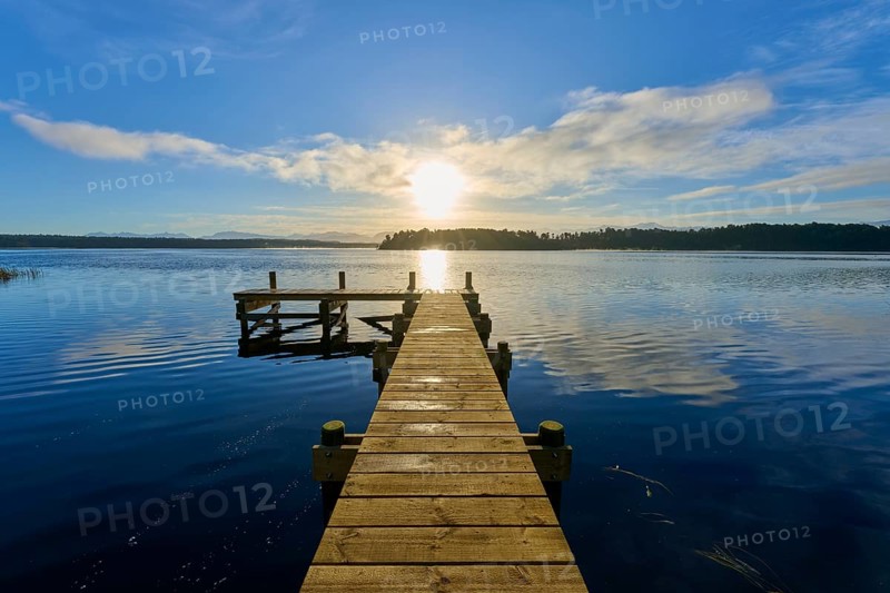 A wooden jetty leads into the calm waters of a lake, the sun is low in the sky, Lake Mahinapua, Ruatapu, South Island, New Zealand