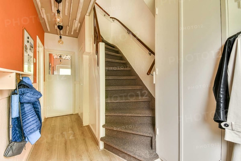 Cozy modern hallway featuring a staircase with carpeted steps leading up. Wooden flooring complements the orange accent wall adorned with hanging lights and coats