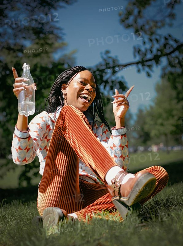 A joyful woman sits in a lush green park, savoring a moment of relaxation with a bottle of water in hand, surrounded by the serenity of nature and the warmth of sunshine