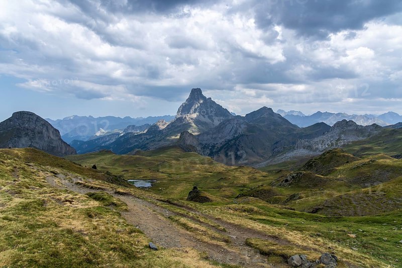Scenic view of the rugged Midi d'Ossau peak surrounded by rolling green hills under a dramatic sky, capturing the serene beauty of the French Pyrenees landscape