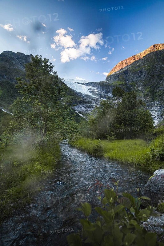 A tranquil stream winds through lush greenery, fed by a glacier nestled in the Norwegian mountains The golden sunlight on the peaks enhances the breathtaking natural beauty