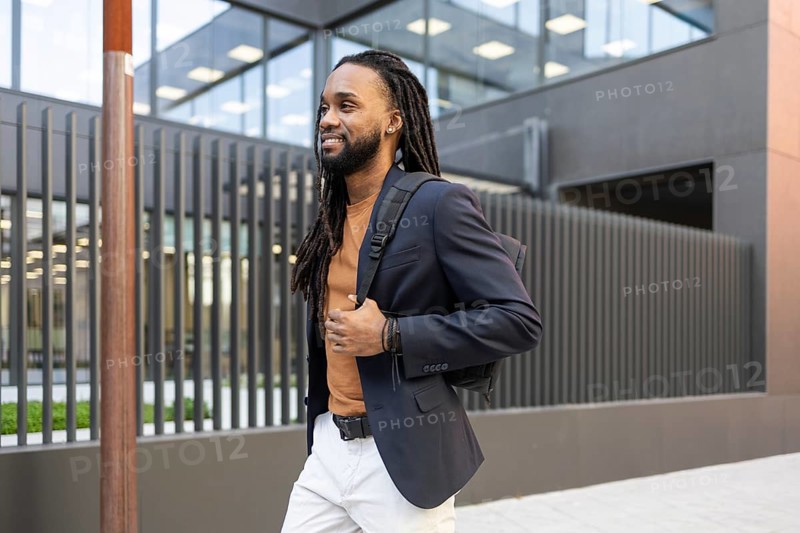 A business man with a backpack walks past a sleek, modern building, exuding style and poise. The contemporary setting complements his professional attire