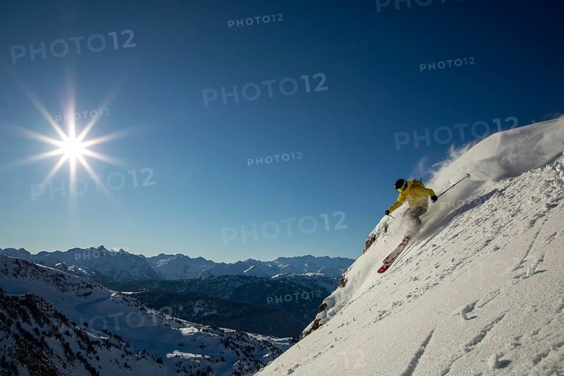 Experience the thrill of freeride skiing on a sunlit, snowy mountain slope A skier in a yellow jacket carves through fresh powder, set against a backdrop of majestic peaks