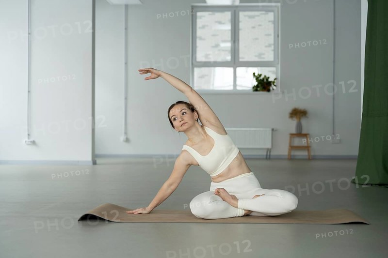 A woman in white active wear practices yoga on a mat in a calm, sunlit studio. The serene indoor space emphasizes focus and wellness, with soft natural lighting enhancing tranquility