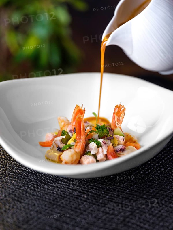 A gourmet dish of seafood featuring shrimp, sliced vegetables, and broth is artfully served in a stylish white bowl. Sophisticated presentation highlights the culinary finesse