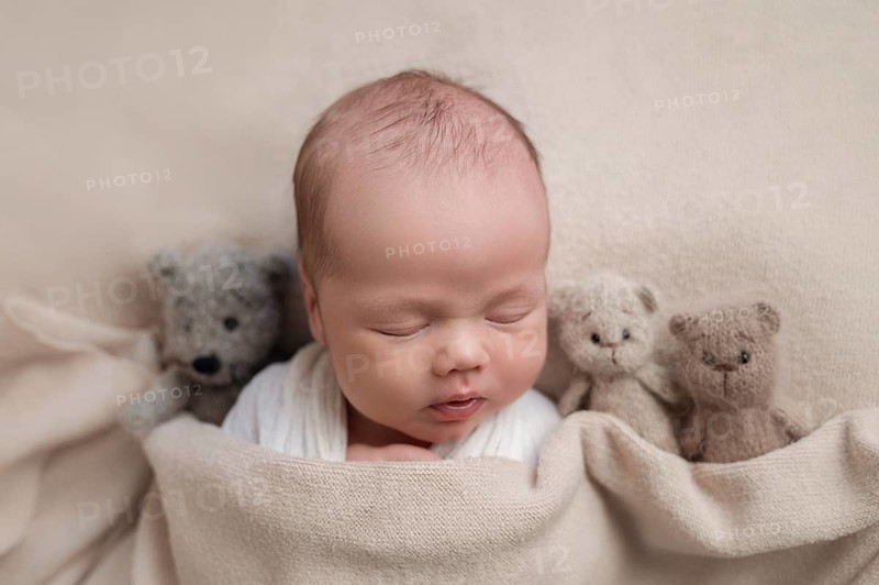 A serene newborn baby sleeps surrounded by three cuddly teddy bears on a soft beige blanket, depicting a warm, comforting scene