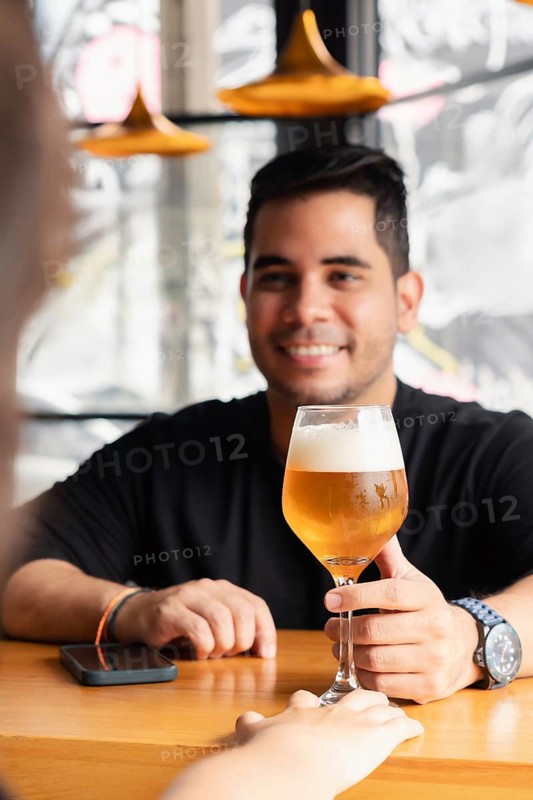 Selective focus on a glass of lager beer with a smiling Latino man in the background receiving the drink at the bar counter. The setting is a modern bar with large windows during daylight hours