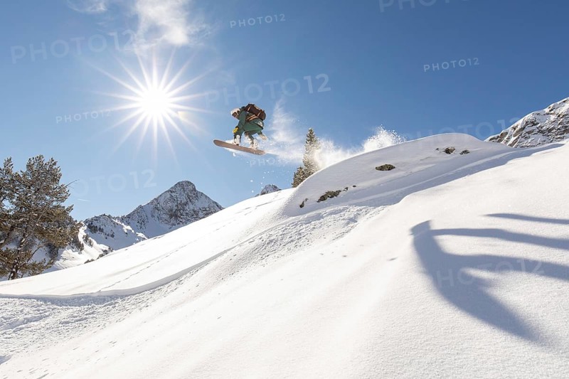 A snowboarder executes an impressive jump on a sunlit, snowy mountain slope, showcasing the thrill of backcountry freeride in fresh powder under a bright blue sky