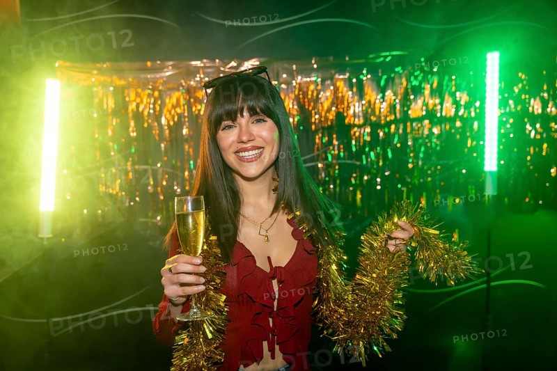 Young smiling woman celebrating during a party, holding a glass of champagne and gold tinsel garland, illuminated by green and yellow neon lights with a festive metallic fringe background