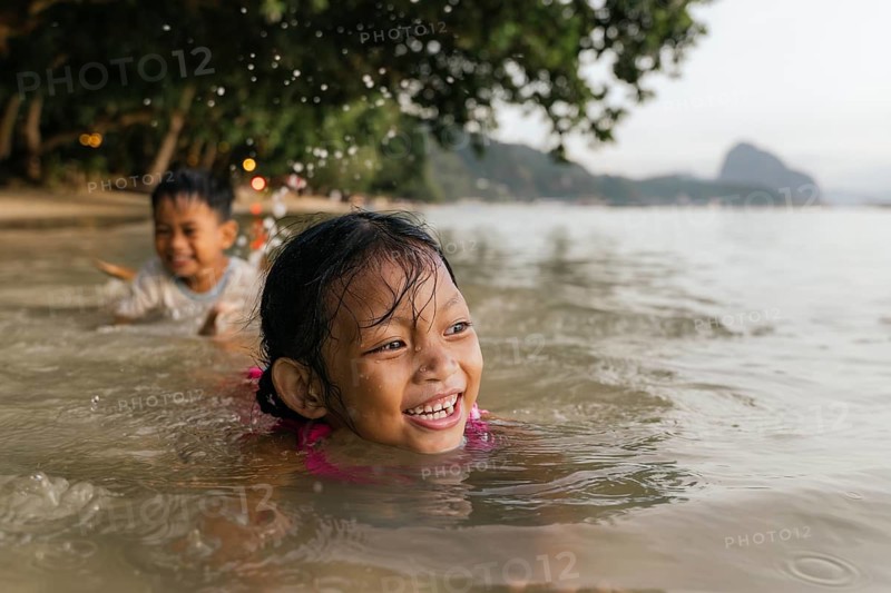 Children happily swim and play in the shallow waters of a picturesque Philippine beach, surrounded by lush greenery and distant hills under a serene sky