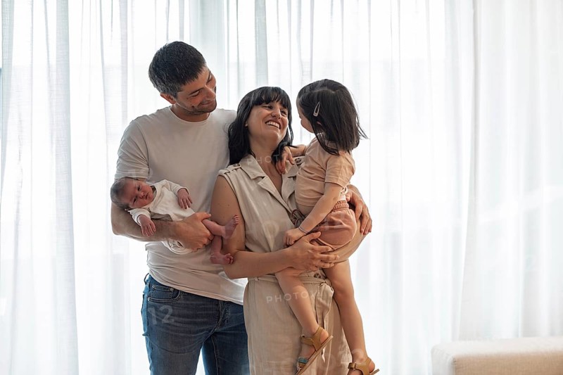 A joyful young family shares a tender moment at home. The image captures a smiling mother and father holding their newborn baby and a sweet big sister in a loving embrace