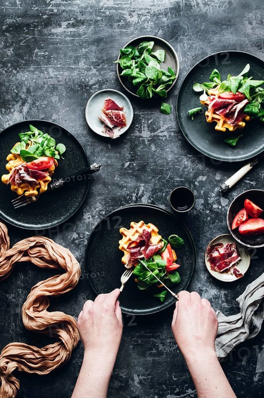 A combination of savory Iberian ham and pumpkin waffles served on dark plates. Garnished with fresh vegetables, this gourmet dish offers an exquisite culinary experience