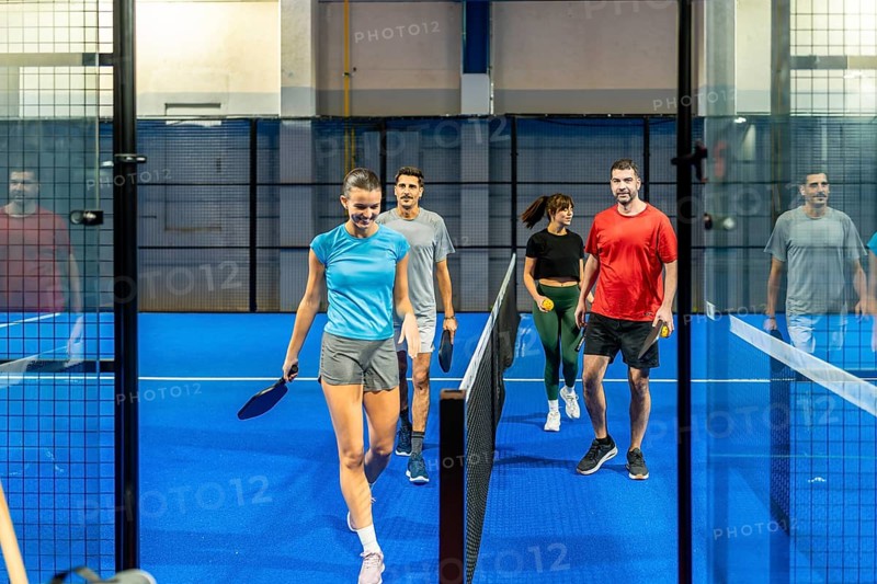 Four friends enter a bright blue pickleball court carrying paddles and balls, smiling and ready to play a casual indoor game focused on fun, fitness and teamwork