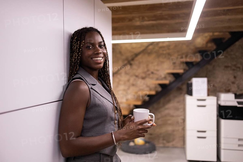 Young African woman with braids stands smiling holding a mug, enjoying a break in a contemporary coworking space. The setting is modern with warm tones and stylish decor