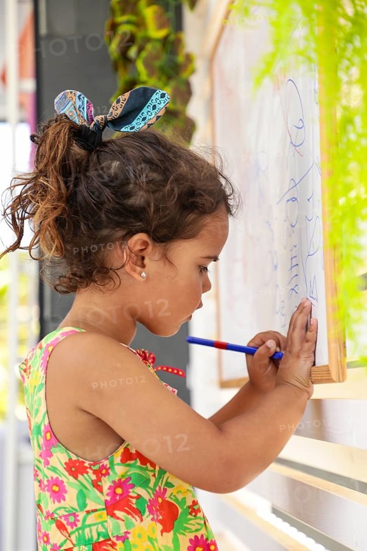 Young girl concentrating as she draws on a whiteboard at home. Her colorful dress and focused expression capture childhood creativity and learning in a warm, family setting