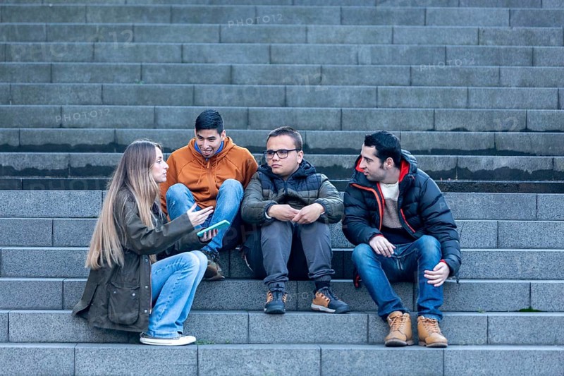 A caregiver sits on outdoor steps with three young men with intellectual disabilities, engaging in a friendly conversation