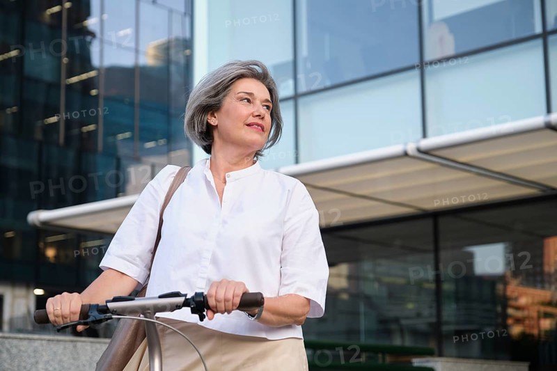 A mature woman with gray hair and a confident smile rides an electric scooter in an urban setting, symbolizing active living and embracing modern technology