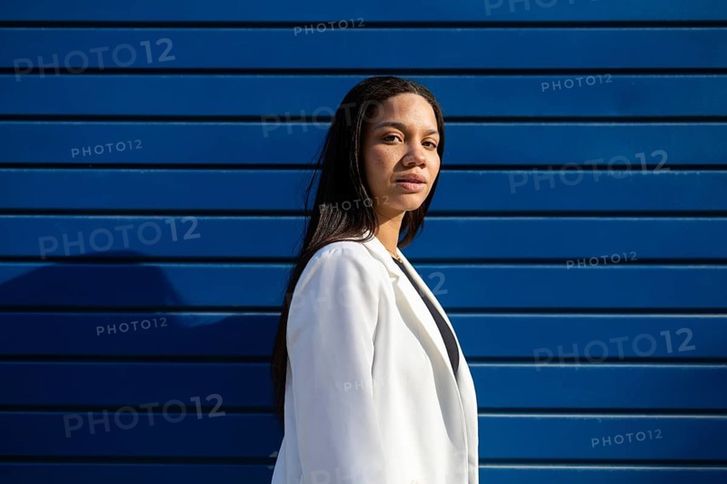 A professional woman in a white blazer stands confidently against a striking blue wall in the sunlight. The image captures a modern business lifestyle theme