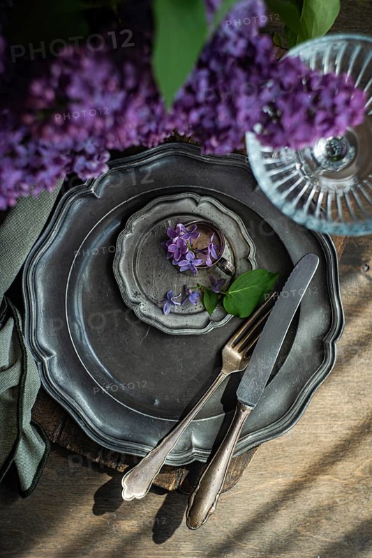 Elegant table setting featuring vintage silverware on tarnished plates, adorned with purple flowers and green leaves. Light streams enhance its rustic charm